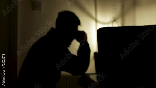 Silhouette of distressed young man sat at table with atmospheric lighting, with his head in hands. Static. photo