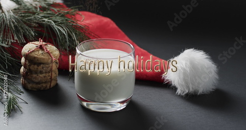 Happy holidays text over christmas hat, cookies and milk on dark grey background