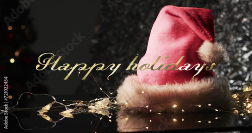Happy holidays text in gold over christmas hat, lights and tree on dark background