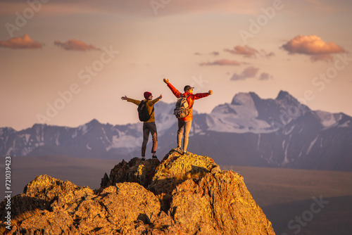 Two happy young tourists with backpacks are standing with open arms at mountain top and enjoys sunset view