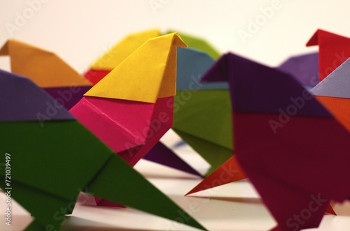 Side angle of a single pink and yellow origami bird standing out while other birds pass by. Part of a series of images of traditional origami birds acting out scenes against a white background.