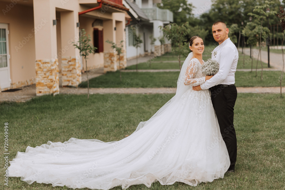 Fototapeta premium A handsome groom embraces his bride in a lush white dress and smiles in a beautiful outdoor setting. Under the open sky. High quality photo. A newlywed couple poses together on a sunny summer day.