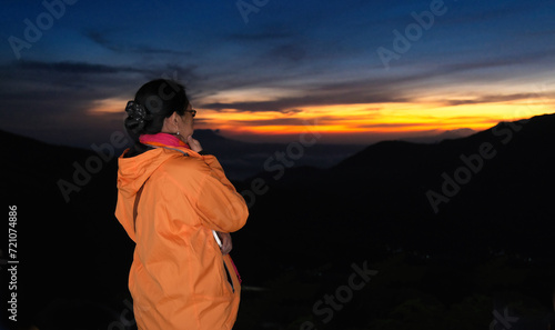 Female mountain climber in orange jacket standing alone to welcome the morning sun reflected on clear sky photo