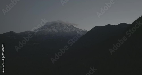 Ungraded wide-angle aerial drone footage of Mount Rainier at the break of dawn as sunrise illuminates the dark, early morning sky above Gifford Pinchot National Forest in Washington State.