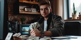 Young, confident and wealthy man is sitting in his studio and holding a wad of money