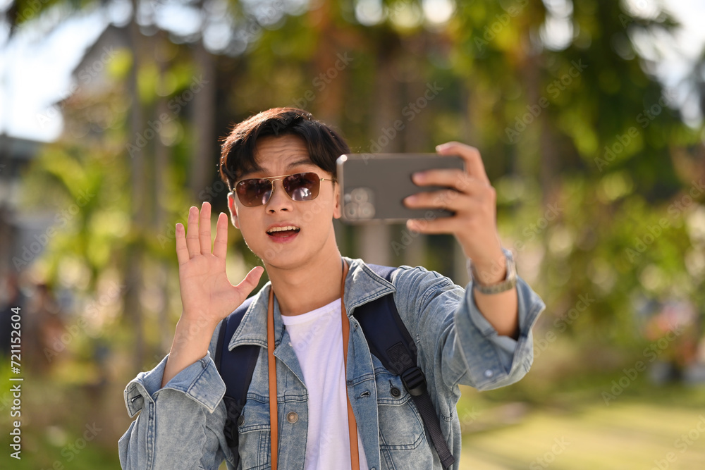 A handsome Asian tourist wears sunglasses waving to the audience who watching the live broadcast on his mobile phone, Travel Influencer and Solo traveler concept A handsome Asian tourist wears sunglasses waving to the audience who watching the live broadcast on his mobile phone, Travel Influencer and Solo traveler concept