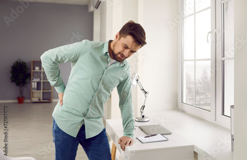 Man suffering from strong back pain. Tired, stressed young man with lumbago backache standing by his working desk, holding his hand on his lower back and making an unhappy face expression