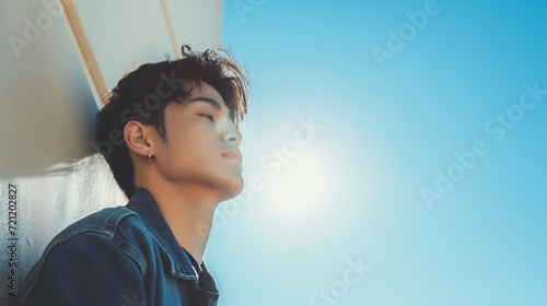Profile headshot of young korean model looking stylish man leaning on the street wall on sunny day with head up tired or posing and clear blue sky copyspace background photo