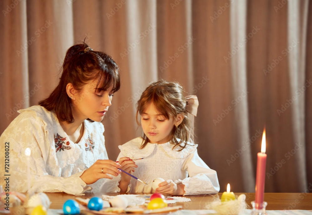 Naklejka premium Cozy moment at home. Beautiful young woman, mother sitting at home with daughter, preparing for Easter, painting eggs. Concept of holiday, Easter, family, motherhood and childhood