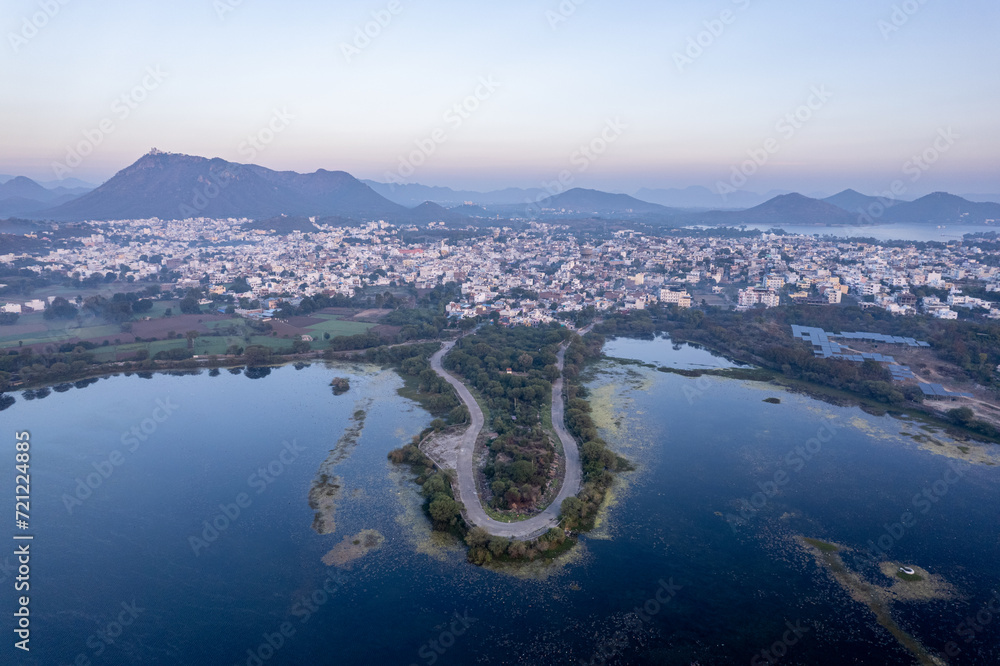 aerial drone shot at dawn dusk with road loop extending into fateh sagar lake with aravalli hills in distance hidden in fog showing cityscape of Udaipur Rajasthan