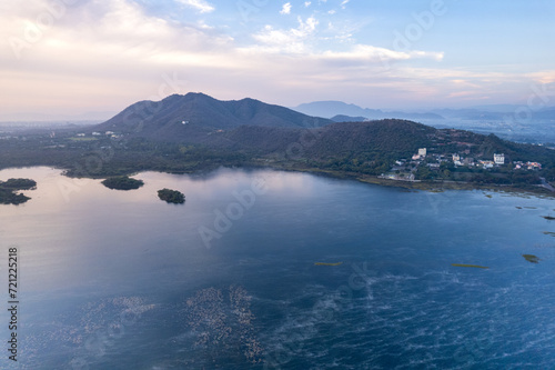 Aerial drone shot dawn dusk sky with aravalli hills with homes, houses at base and blue weed covered waters of Pichola Fateh Sagar lake in Udaipur, Nainital
