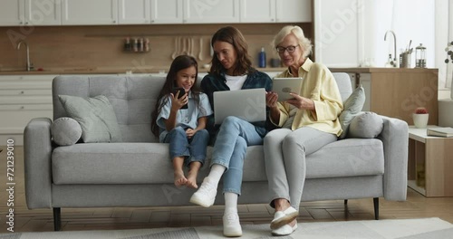 Cute child girl, young mother and grandmother discussing online content on gadgets, sitting on sofa with cellphone, laptop, tablet, using Internet application, service, communication photo