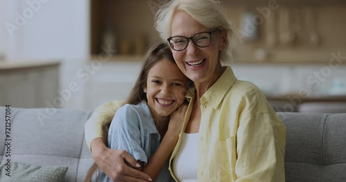 Cheerful cute granddaughter kid and loving grandma hugging on comfortable couch, smiling at camera, laughing, turning look at each other, enjoying funny family leisure, close friendship photo