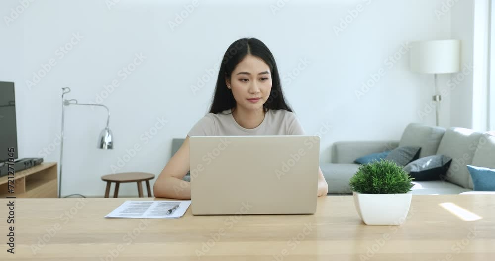 Serious busy young Asian entrepreneur woman typing on laptop, sitting at large workplace table, working on business project at home, reading, watching content, touching chin, thinking Serious busy young Asian entrepreneur woman typing on laptop, sitting at large workplace table, working on business project at home, reading, watching content, touching chin, thinking