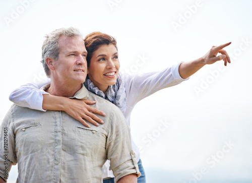 Happy, mature couple and pointing on holiday with travel to beach and embrace with love on adventure in summer. Vacation, sightseeing and woman hug man in nature on blue sky with mock up space