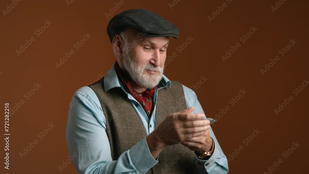 A man of seasoned years expresses curiosity as he counts a handful of bills, attired in a classic flat cap and tweed vest, against a warm brown background. High quality 4k footage