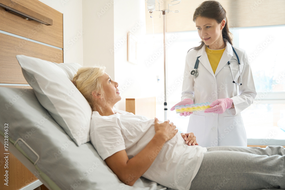 Doctor showing pill box to pregnant woman on medical bed in maternity clinic Doctor showing pill box to pregnant woman on medical bed in maternity clinic