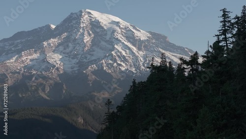 Soaring aerial view of Mount Rainier's snow-capped peak, standing grand against the blue sky, framed by the evergreen forests of Washington State.