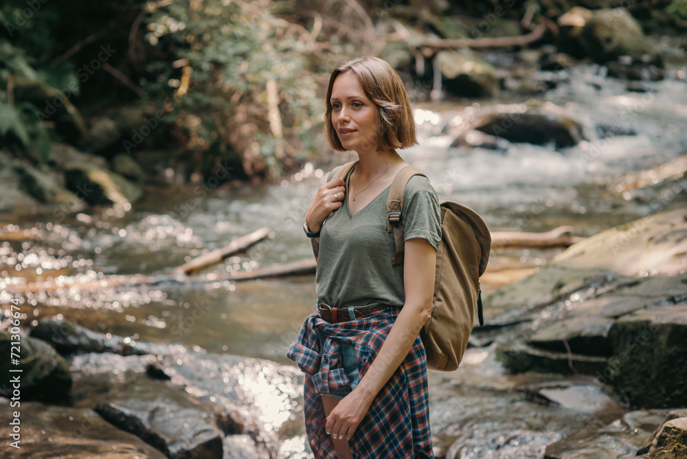 Attractive young female traveler with backpack standing by the mountain river and enjoying views