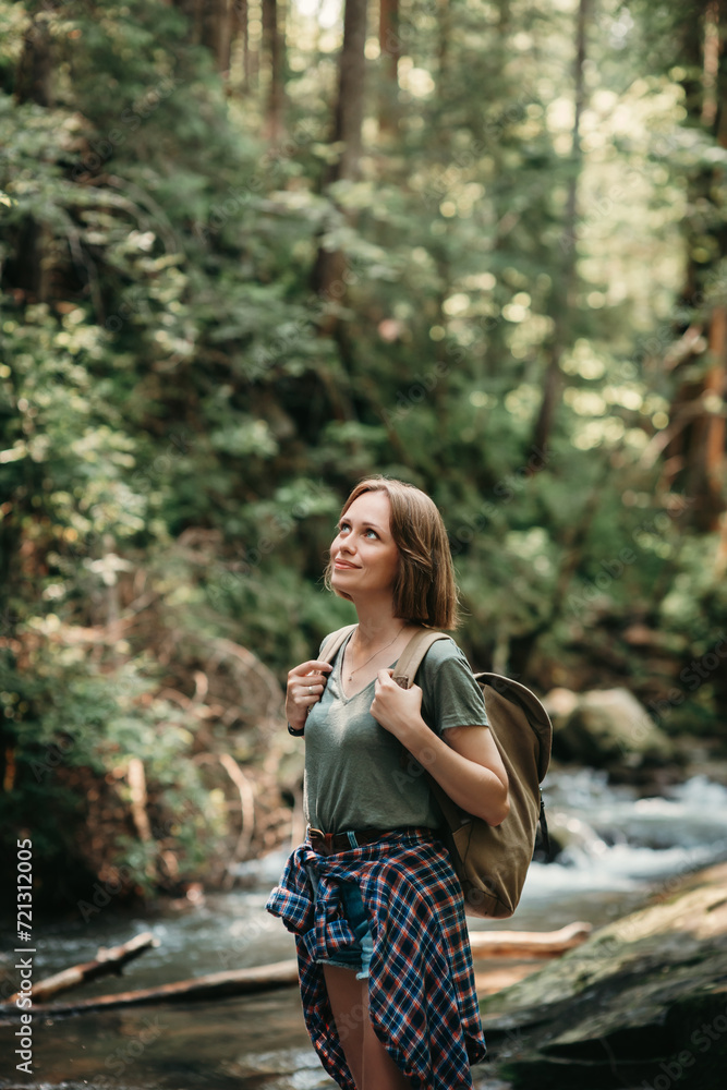 Smiling young female traveler with backpack standing by the mountain river and enjoying views Smiling young female traveler with backpack standing by the mountain river and enjoying views