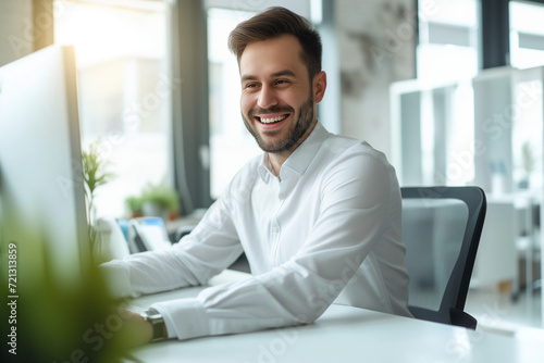 Portrait of a young man with a beard, wearing smart casual attire, displaying a warm, friendly smile as he works at his computer in a modern, naturally lit office space. His appearance is neat and pro © Duncan