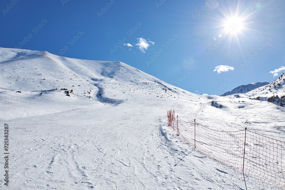 Empty piste at ski resort, mountain slope. Red mesh fence marking the dangerous area and the border of the route. Winter mountains landscape at sunny day. No people. Snowboarding track. Active travel Empty piste at ski resort, mountain slope. Red mesh fence marking the dangerous area and the border of the route. Winter mountains landscape at sunny day. No people. Snowboarding track. Active travel