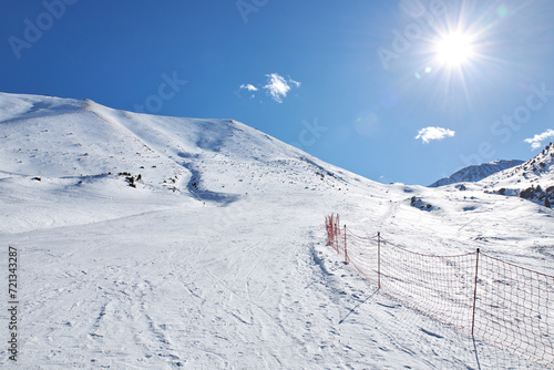 Empty piste at ski resort, mountain slope. Red mesh fence marking the dangerous area and the border of the route. Winter mountains landscape at sunny day. No people. Snowboarding track. Active travel