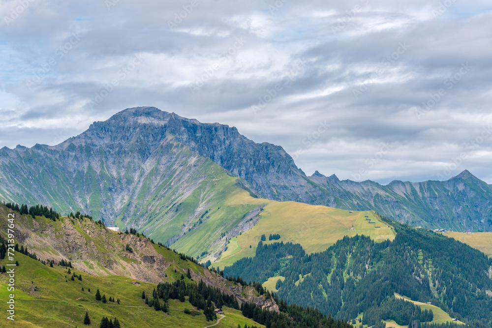 Panoramic view of mountain ridges and slopes with green pastures in summer and cloudy sky in the background. A view to Gsür 2708 m., a mountain in the Bernese Alps, Adelboden, Switzerland.