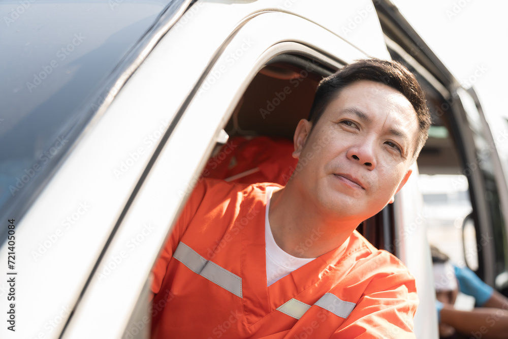 Asian male ambulance staff member in his ambulance car. He is wearing ambulance uniform of paramedics during sitting with paramedics giving injured person first aid background Asian male ambulance staff member in his ambulance car. He is wearing ambulance uniform of paramedics during sitting with paramedics giving injured person first aid background