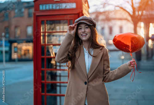 young happy woman with a heart-shaped balloon falling a love, having a fun day, walking against the red phone box in English city Spring is in the air Lifestyle, tourism, valentines day concept