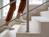 Woman in white sneakers and khaki trousers goes upstairs to her apartment. White staircase in apartment building. Casual outfit, urban fashion. Physical exercises.