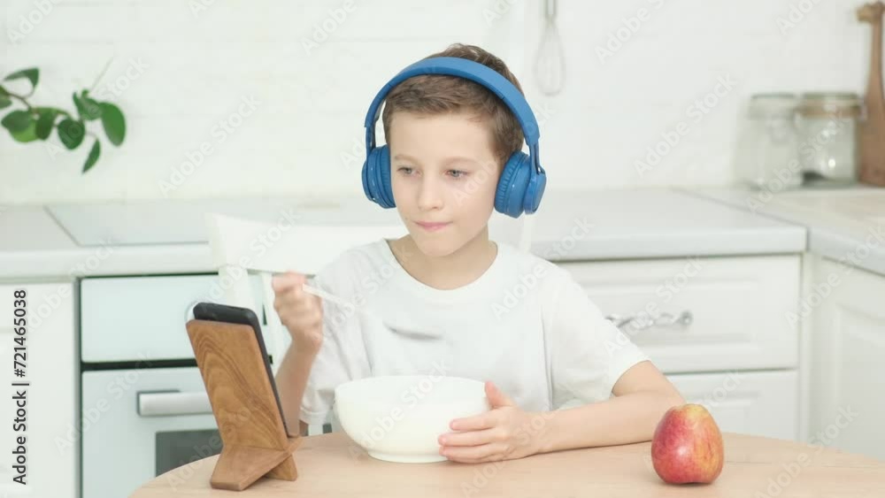 Child boy is watching video and wearing headphones on round wooden table kitchen, wearing headphone in white table Child boy is watching video and wearing headphones on round wooden table kitchen, wearing headphone in white table