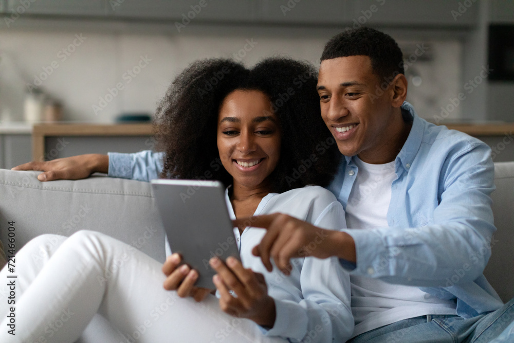 Happy black couple using digital tablet while relaxing on sofa in living room, shopping online, browsing internet or checking new application Happy black couple using digital tablet while relaxing on sofa in living room, shopping online, browsing internet or checking new application