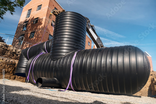 Attenuation tank made of big diameter plastic pipe delivered on construction site, offloaded and moved by an excavator into required position for assembling by builders photo