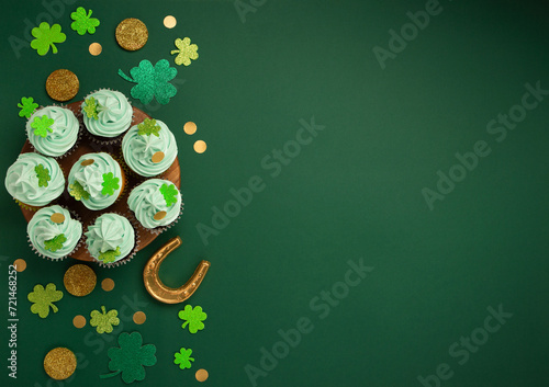 St. Patrick's Day vanilla and chocolate cupcakes with green frosting and  shiny clover decorations on green paper background. Irish holiday dessert concept. Top view, copy space.