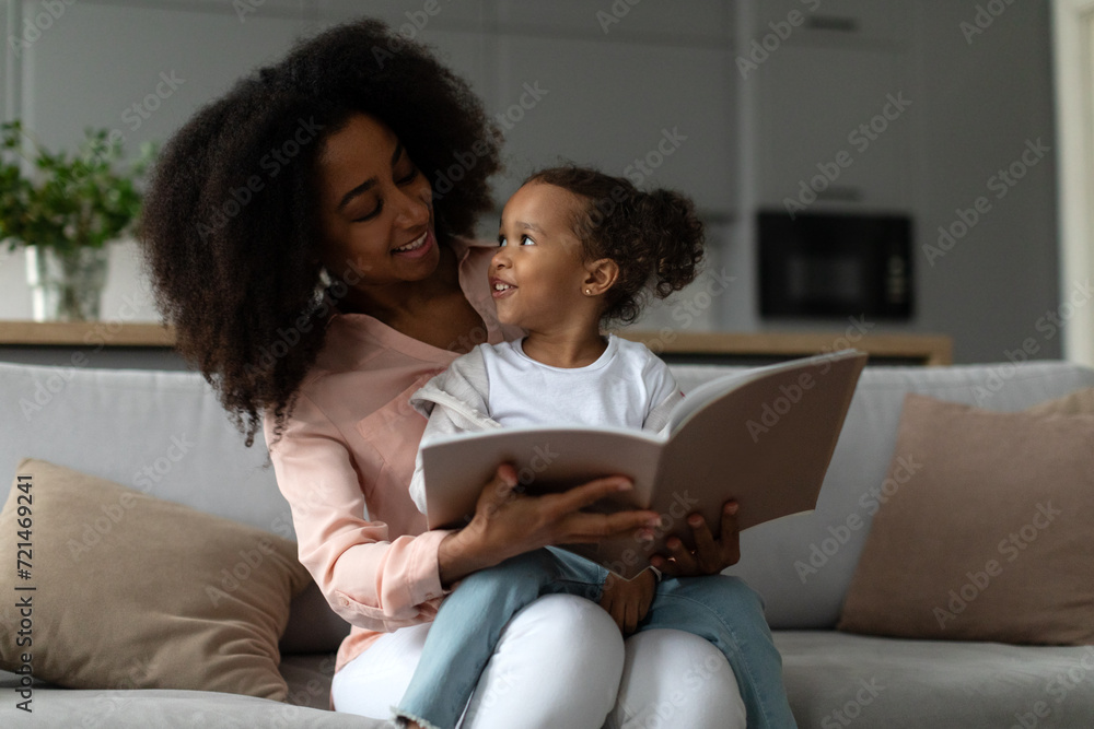 Loving black mother reading book with her little daughter, sitting together on sofa at home, spending time together in living room