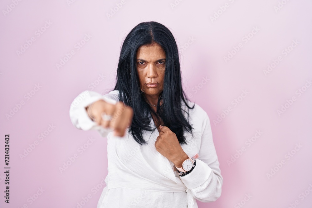 Mature hispanic woman standing over pink background punching fist to fight, aggressive and angry attack, threat and violence Mature hispanic woman standing over pink background punching fist to fight, aggressive and angry attack, threat and violence