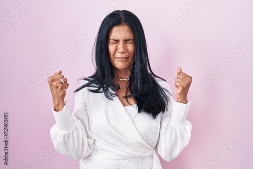 Mature hispanic woman standing over pink background excited for success with arms raised and eyes closed celebrating victory smiling. winner concept.
