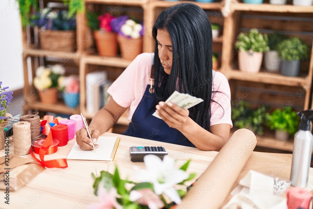 Middle age hispanic woman florist counting dollars writing on notebook at florist