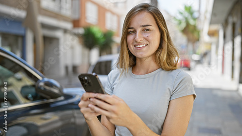 Cheerful young blonde woman happily touching her smartphone, engaged in a digital conversation on a sunny city street