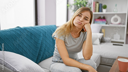 Attractive young blonde woman sitting with a serious expression, contemplating her problems on a sofa at home