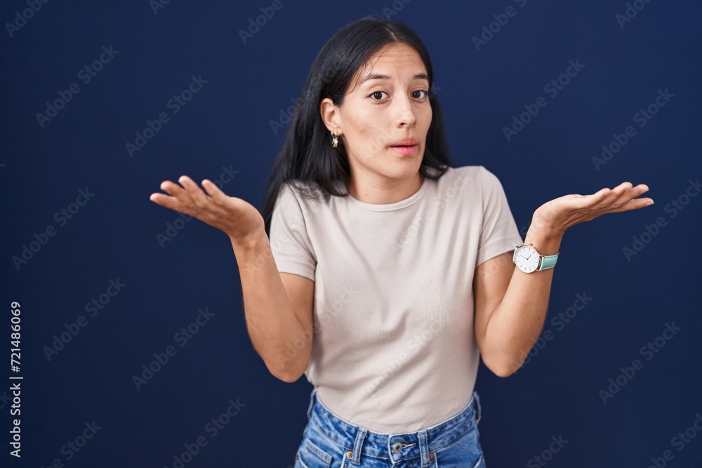 Young hispanic woman standing over blue background clueless and confused expression with arms and hands raised. doubt concept. Young hispanic woman standing over blue background clueless and confused expression with arms and hands raised. doubt concept.