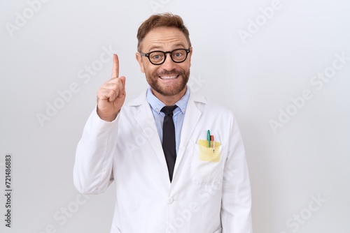Middle age doctor man with beard wearing white coat showing and pointing up with finger number one while smiling confident and happy.