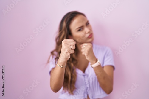 Young hispanic woman standing over pink background ready to fight with fist defense gesture, angry and upset face, afraid of problem