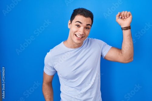 Young hispanic man standing over blue background dancing happy and cheerful, smiling moving casual and confident listening to music