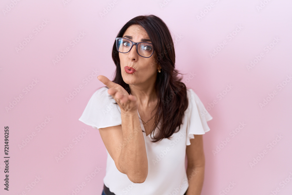 Middle age hispanic woman wearing casual white t shirt and glasses looking at the camera blowing a kiss with hand on air being lovely and sexy. love expression. Middle age hispanic woman wearing casual white t shirt and glasses looking at the camera blowing a kiss with hand on air being lovely and sexy. love expression.