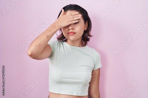 Hispanic young woman standing over pink background covering eyes with hand, looking serious and sad. sightless, hiding and rejection concept