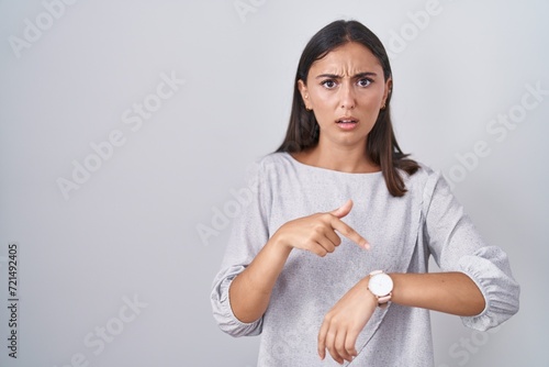 Young hispanic woman standing over white background in hurry pointing to watch time, impatience, upset and angry for deadline delay