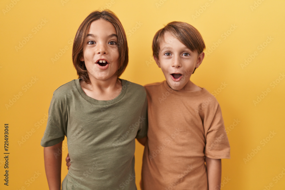 Adorable boys hugging in astonishment, open-mouthed disbelief reaction expressing funny fear and wonder, standing against yellow isolated background - obrazy, fototapety, plakaty 