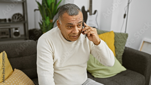 A middle-aged hispanic man talking on a smartphone while sitting on a sofa in a modern living room.